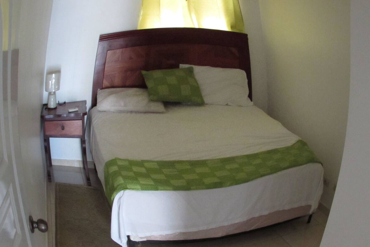 A neatly made bed with a wooden headboard, a bedside table with a lamp, and a green checkered throw blanket.