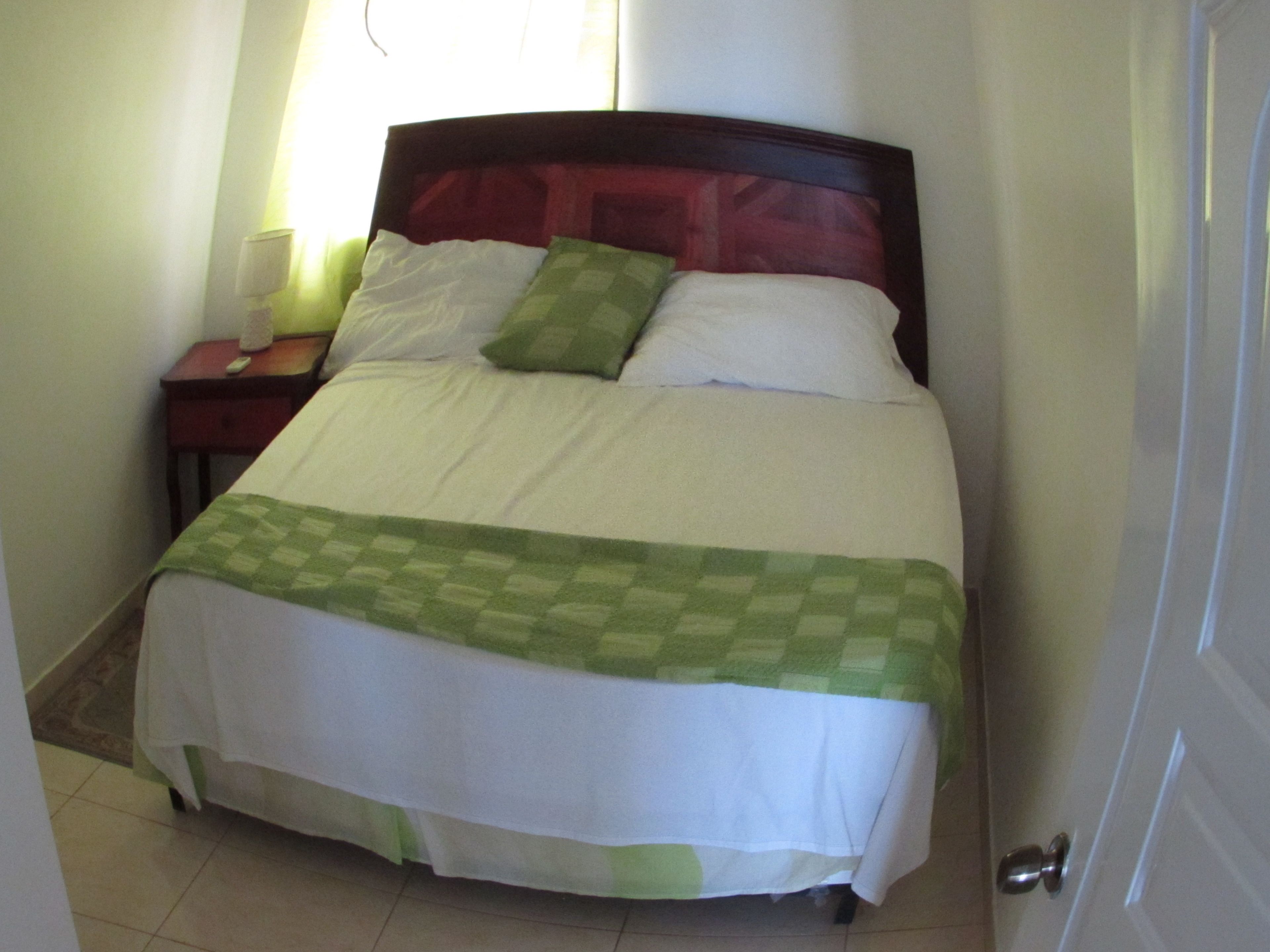 Una cama bien hecha con un edredón verde a cuadros y almohadas blancas, una mesita de noche con una lámpara y una puerta a la derecha.