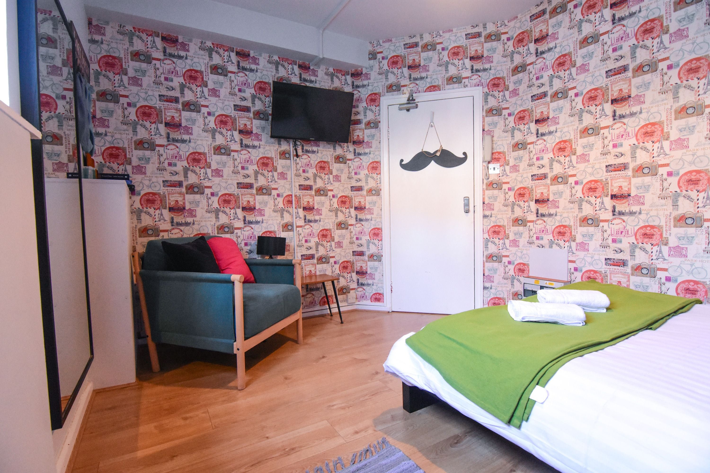 A room with patterned wallpaper, a bed with a green blanket, a blue sofa with a red pillow, a television mounted on the wall, and a door with a mustache decoration.