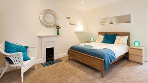 A bedroom with a wooden bed, white wicker chair, and a fireplace. - Monopoly Accommodations (London)