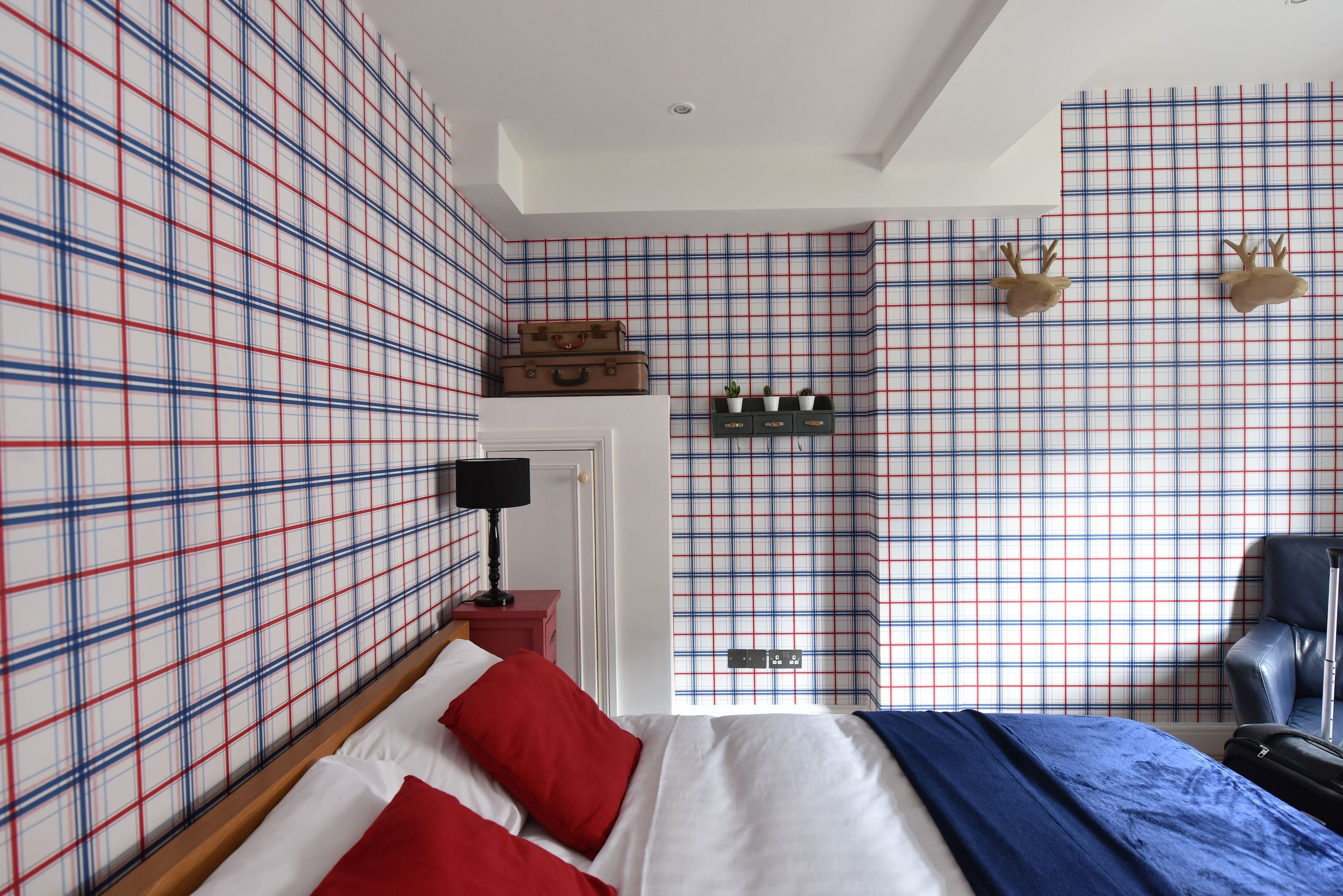 A bedroom with a plaid wallpaper, a bed with red and white bedding, a nightstand with a lamp, a suitcase, and a chair.