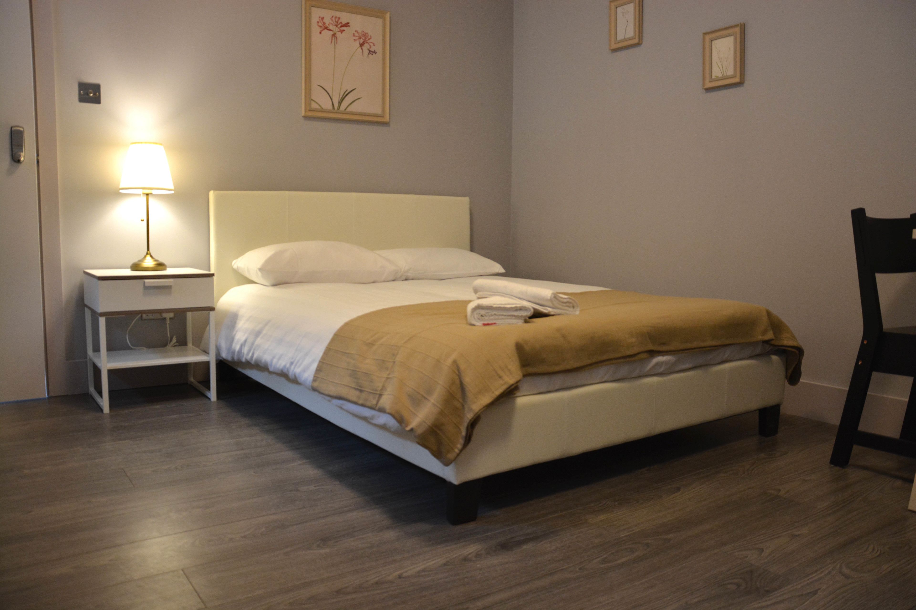 A neatly made bed with a white headboard and a brown blanket, a bedside table with a lamp, and a chair to the right.