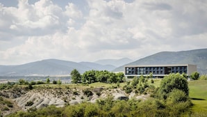 Ein modernes Gebäude mit flachem Dach und großen Fenstern, umgeben von Grünflächen und Bergen im Hintergrund.