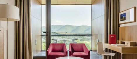 Una habitación de hotel moderna con un ventanal grande, dos sillones rojos, una pequeña mesa redonda, un escritorio con una carpeta roja y una cama.