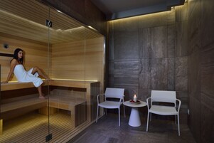 A modern spa with wooden benches, a glass sauna, and two white chairs with a small table and candle.