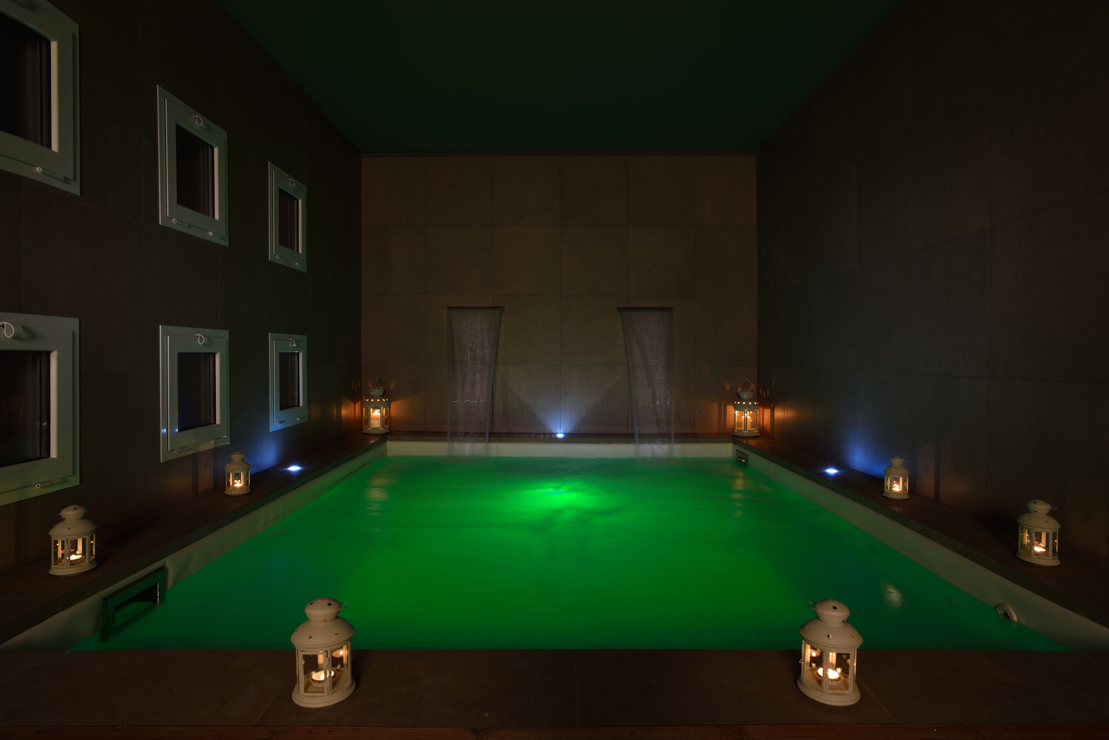 A dimly lit indoor swimming pool with green lighting, surrounded by walls with framed windows and wall-mounted lanterns.