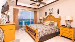 A bedroom with a bed, wooden nightstands, a ceiling fan, a TV, and a view of the ocean. - Villa Del Playa Penthouse 5 (Roatan)