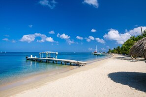 A beach with a wooden pier extending into the water, clear blue skies, and a few boats in the distance. - Villa Del Playa Penthouse 5 (Roatan)