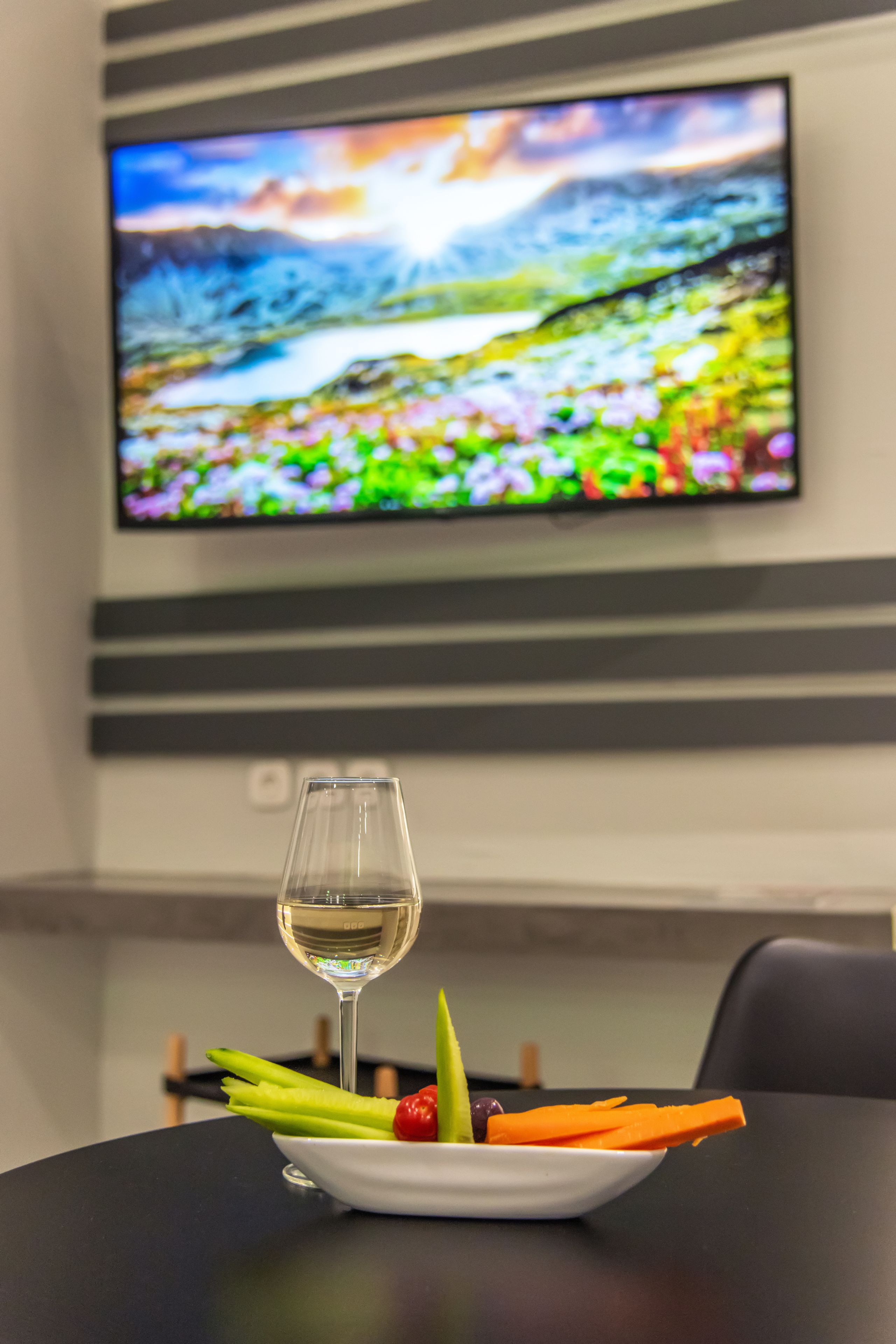 A dining table with a white plate of vegetables and a glass of white wine, set against a backdrop of a television displaying a colorful landscape.
