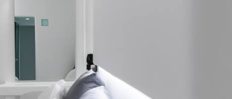 Habitación de hotel moderna con una cama blanca, almohadas grises, una mesita de noche blanca y un aire acondicionado instalado en la pared.