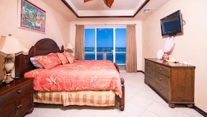 A bedroom with a bed, dresser, TV, and a view of the ocean. - Villa Del Playa Penthouse 6 (Roatan)