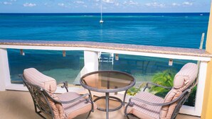 A balcony with two cushioned chairs and a glass table overlooking a clear blue sea with a sailboat in the distance. - Villa Del Playa Penthouse 6 (Roatan)