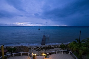 Blick auf einen Strand mit einem Pier, der ins Meer führt, Palmen und einer Struktur mit einem Reetdach.