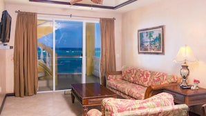 A room with a sofa, a table, a lamp, a painting, and a view of the ocean through a sliding door. - Villa Del Playa Penthouse 6 (Roatan)