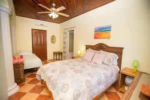 A bedroom with a wooden ceiling, a bed with patterned bedding, a bedside table with a lamp, and a door.