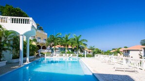 Ein Swimmingpool mit weißen Geländern, Palmen und Gebäuden mit Balkonen im Hintergrund.