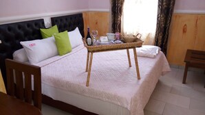A neatly made bed with a headboard, pillows, a bedside table with a tray, and a chair. - Liza Hôtel (Yaoundé)