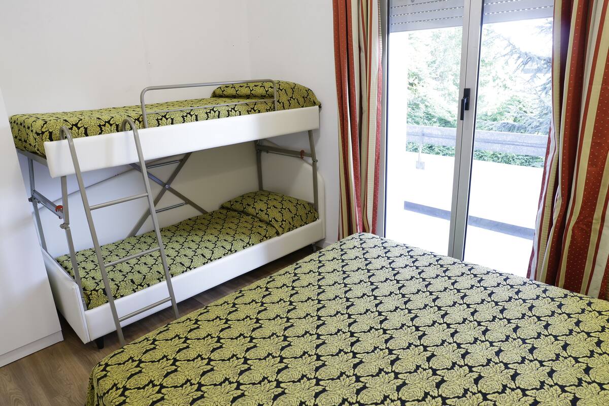 A bunk bed with a patterned bedspread, a window with curtains, and a view of greenery outside.