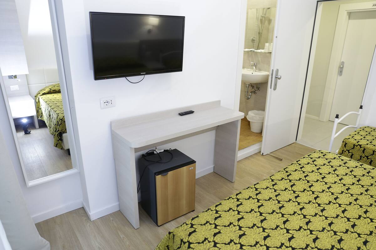 A hotel room with a bed, a desk with a TV, a small storage unit, and a bathroom visible through an open door.