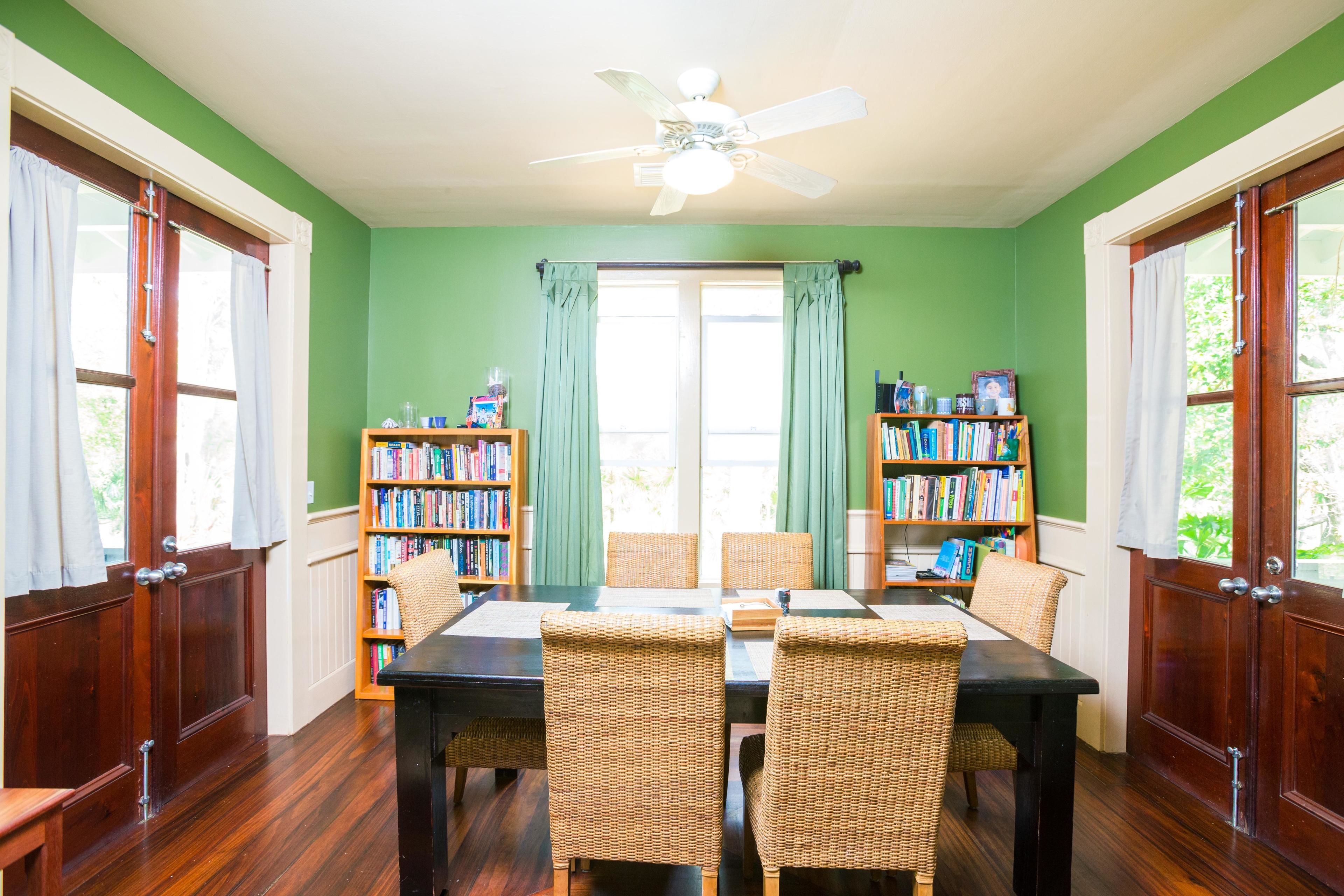 A dining room with a wooden table, four wicker chairs, two bookshelves, and a ceiling fan.