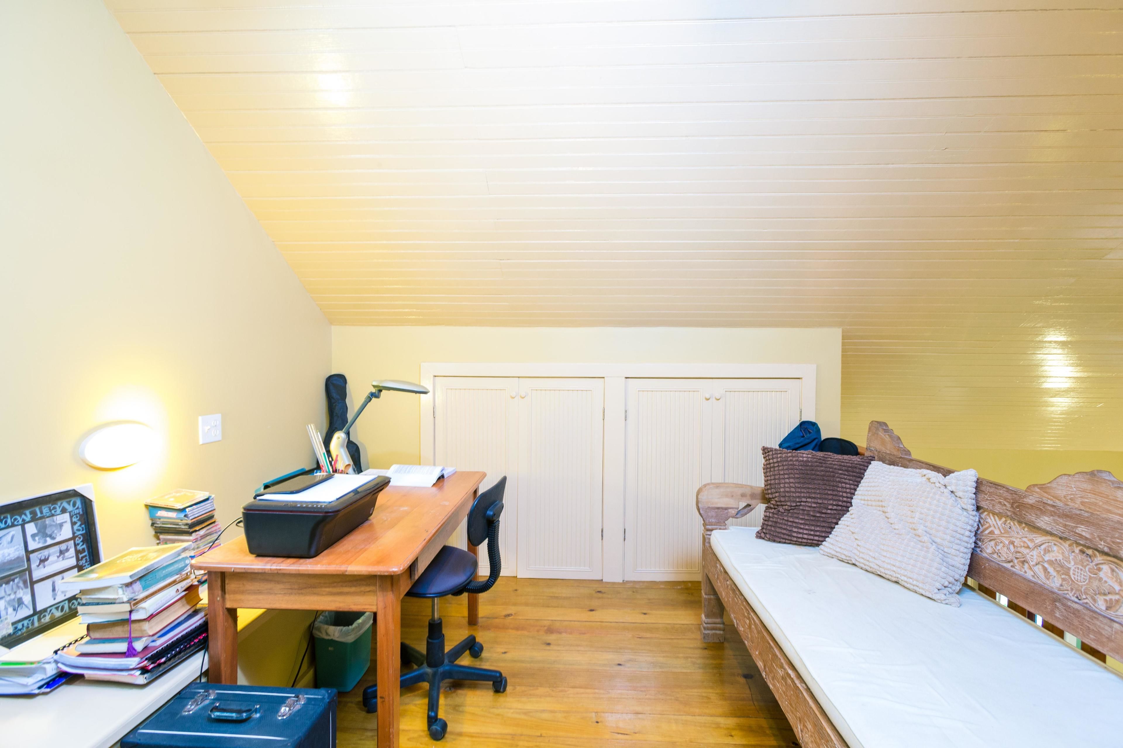 Una habitación pequeña y acogedora con un escritorio de madera, una cama con almohadas y una estantería con libros y una lámpara.