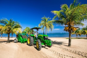 A green tractor on a sandy beach with palm trees and the ocean in the background.