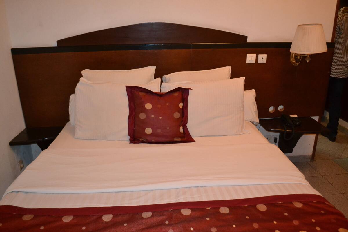 A neatly made bed with white linens and a red polka dot blanket. There is a wooden headboard and a bedside table with a lamp.