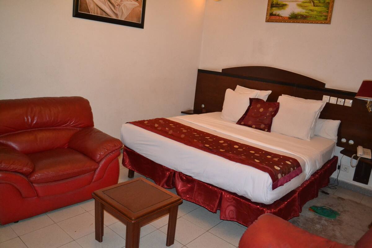 A hotel room with a bed, a red leather sofa, a small wooden table, and a nightstand with a lamp.