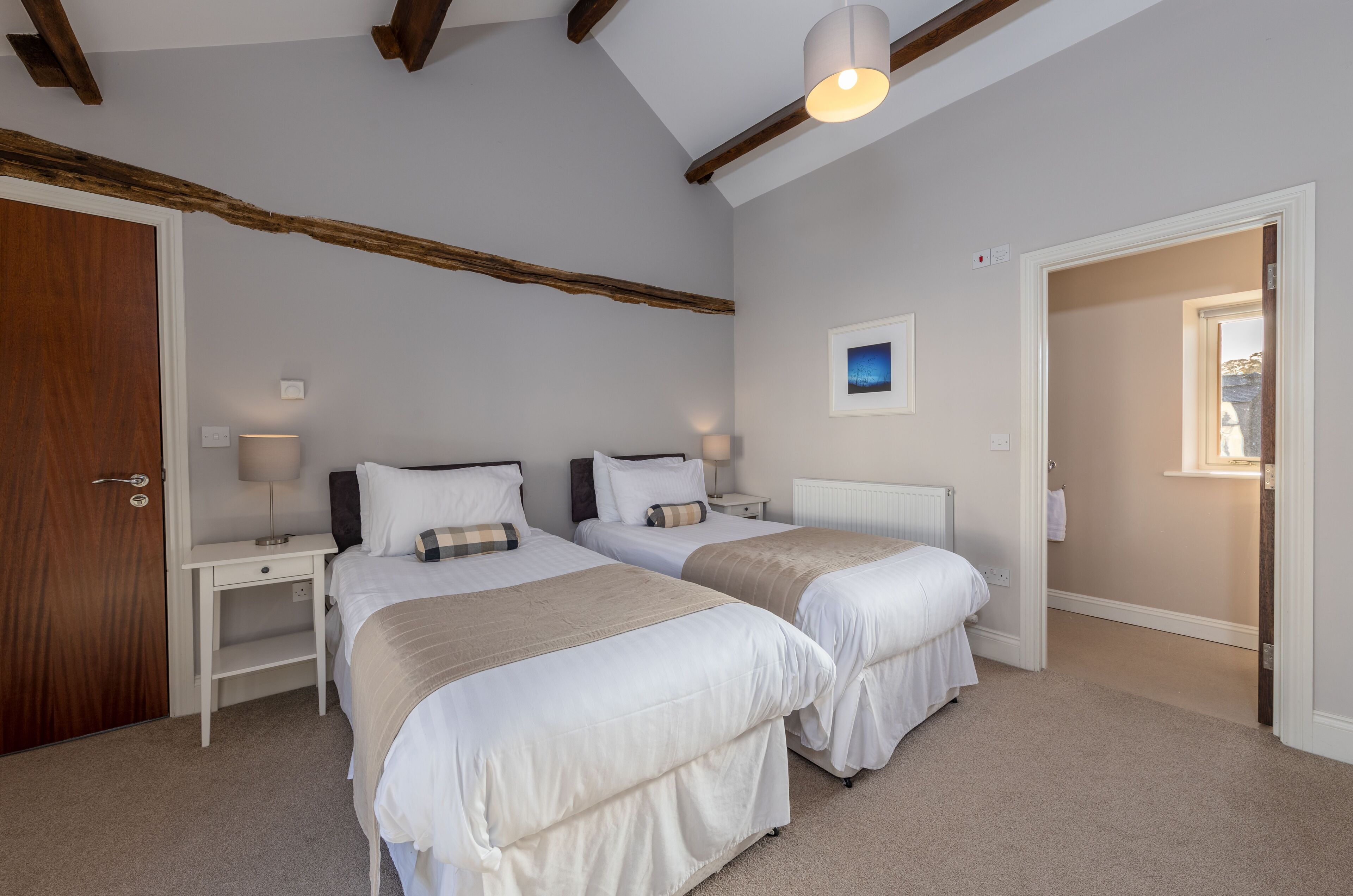 A bedroom with two beds, a wooden ceiling beam, a nightstand with a lamp, and a framed picture on the wall.
