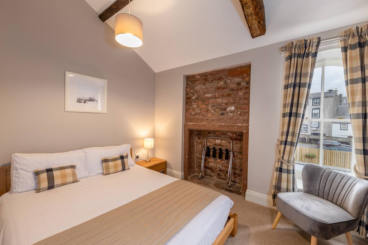 A bedroom with a large bed, a fireplace, a chair, and a window with curtains.