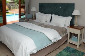 A bedroom with a large bed, two bedside tables with lamps, a view of a swimming pool, and a small side table with a box. - Oleander Guest House (Kimberley)