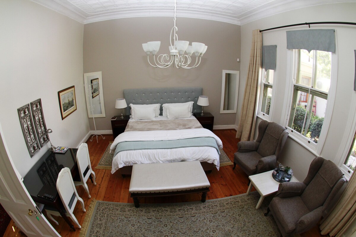 A bedroom with a large bed, two bedside tables, a chandelier, a sofa, and a window with curtains.
