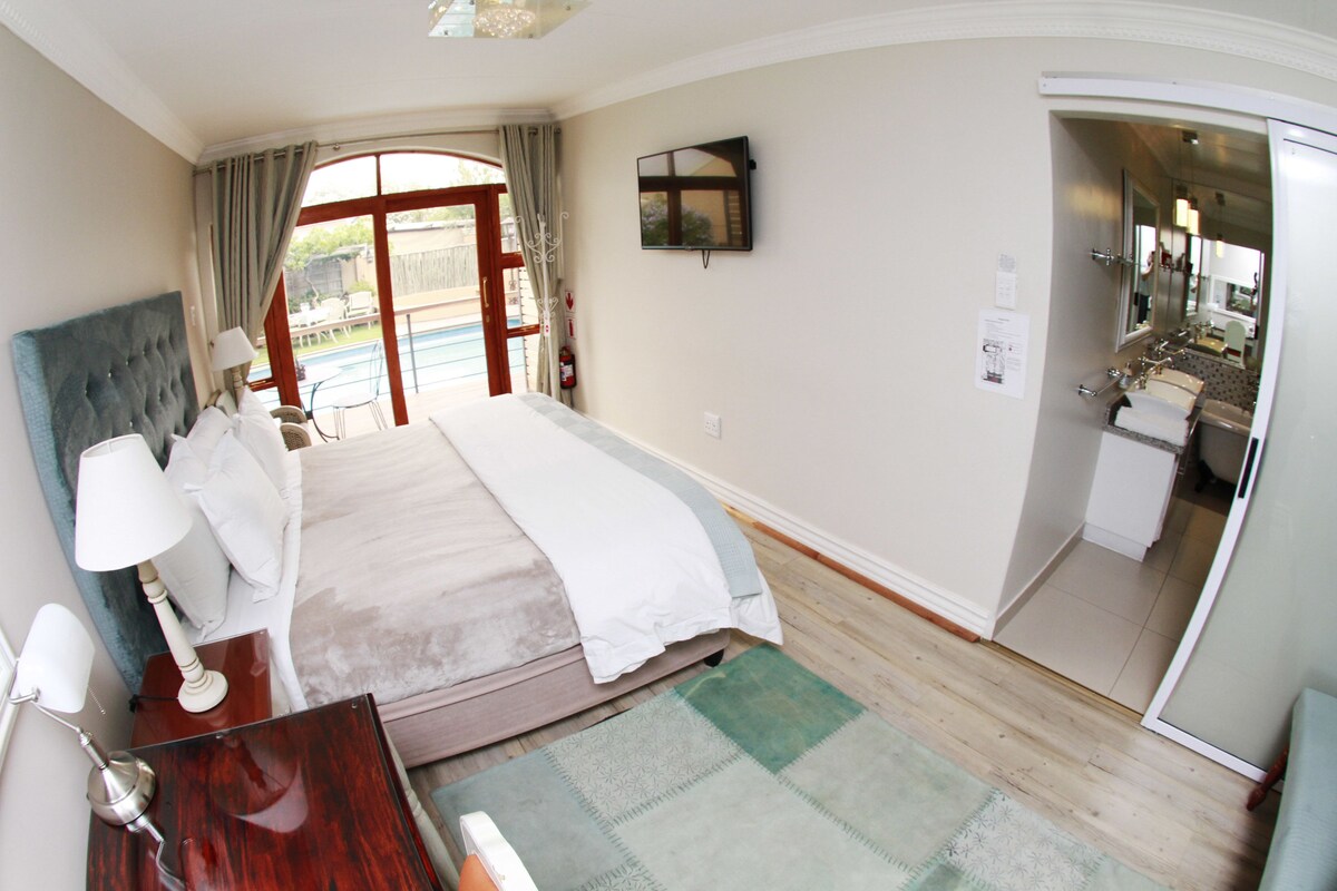 A bedroom with a large bed, a bedside table with a lamp, a view of the outside through a glass door, and a bathroom visible through an open door.
