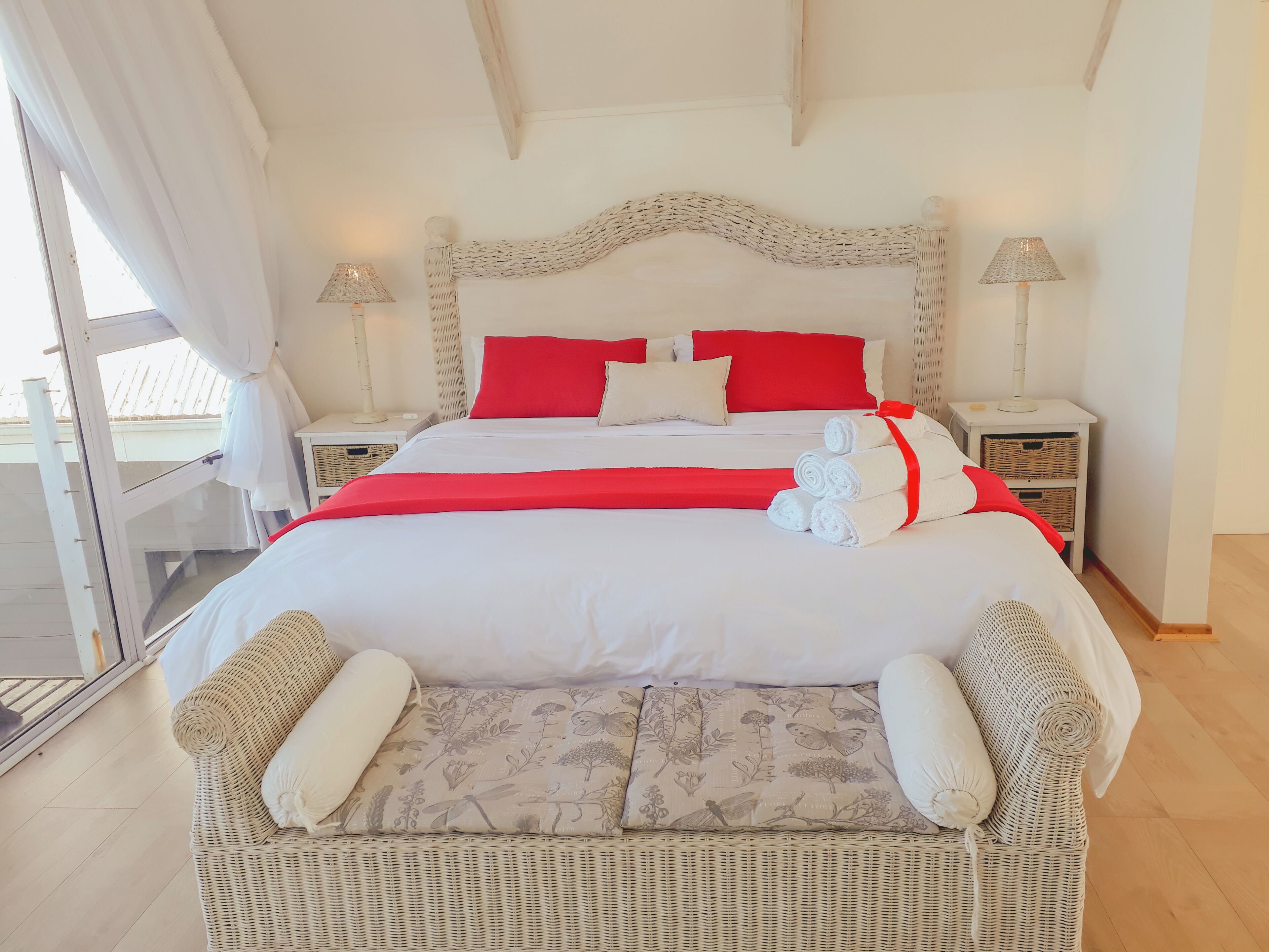 A bed with white and red bedding, two nightstands with lamps, a wicker chair with cushions, and a view of the outdoors through a glass door.