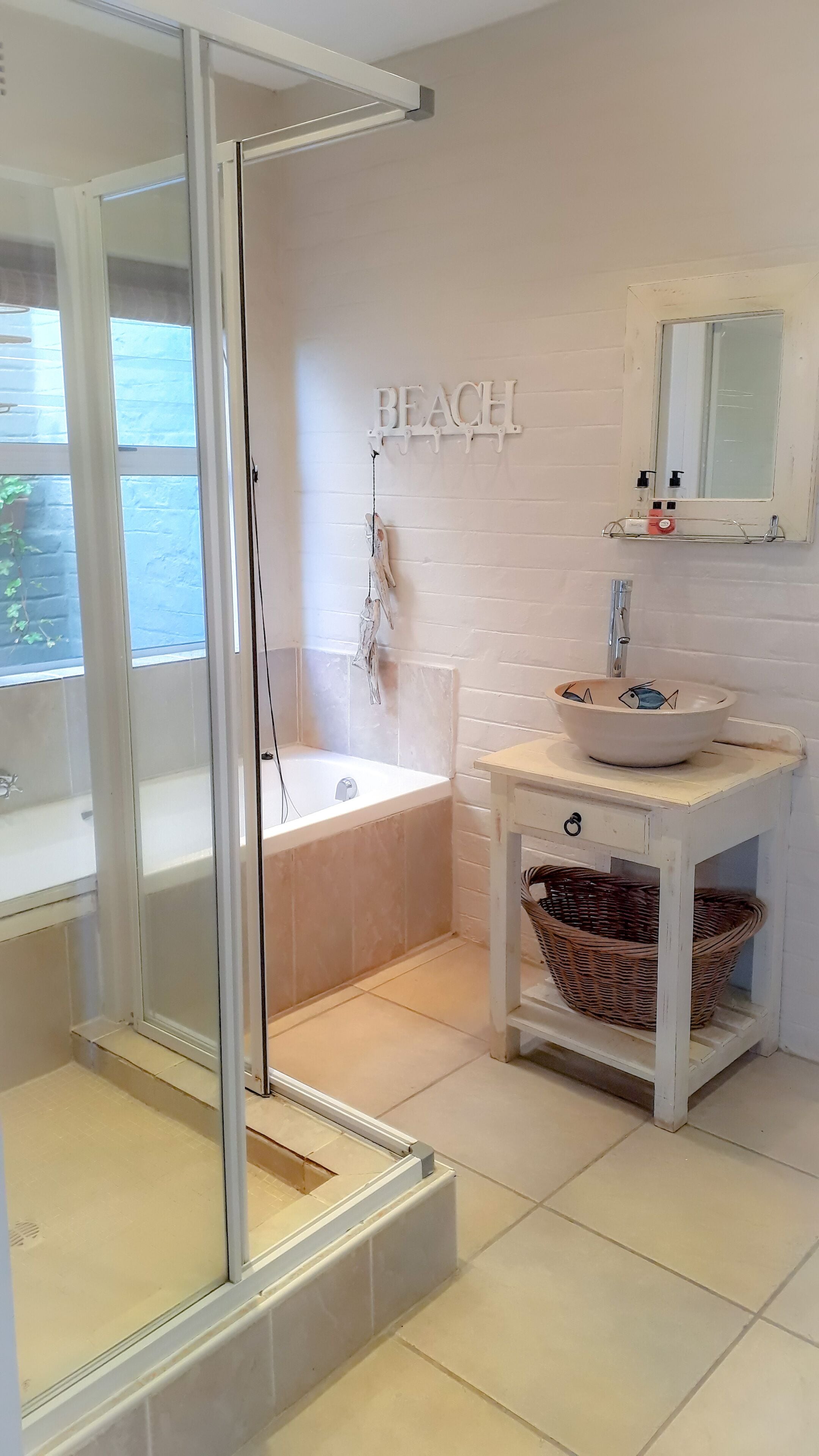 Une salle de bain avec une douche à parois de verre, une baignoire, un lavabo agrémenté d’un récipient décoratif, un miroir et un panneau mural indiquant le mot « PLAGE ».