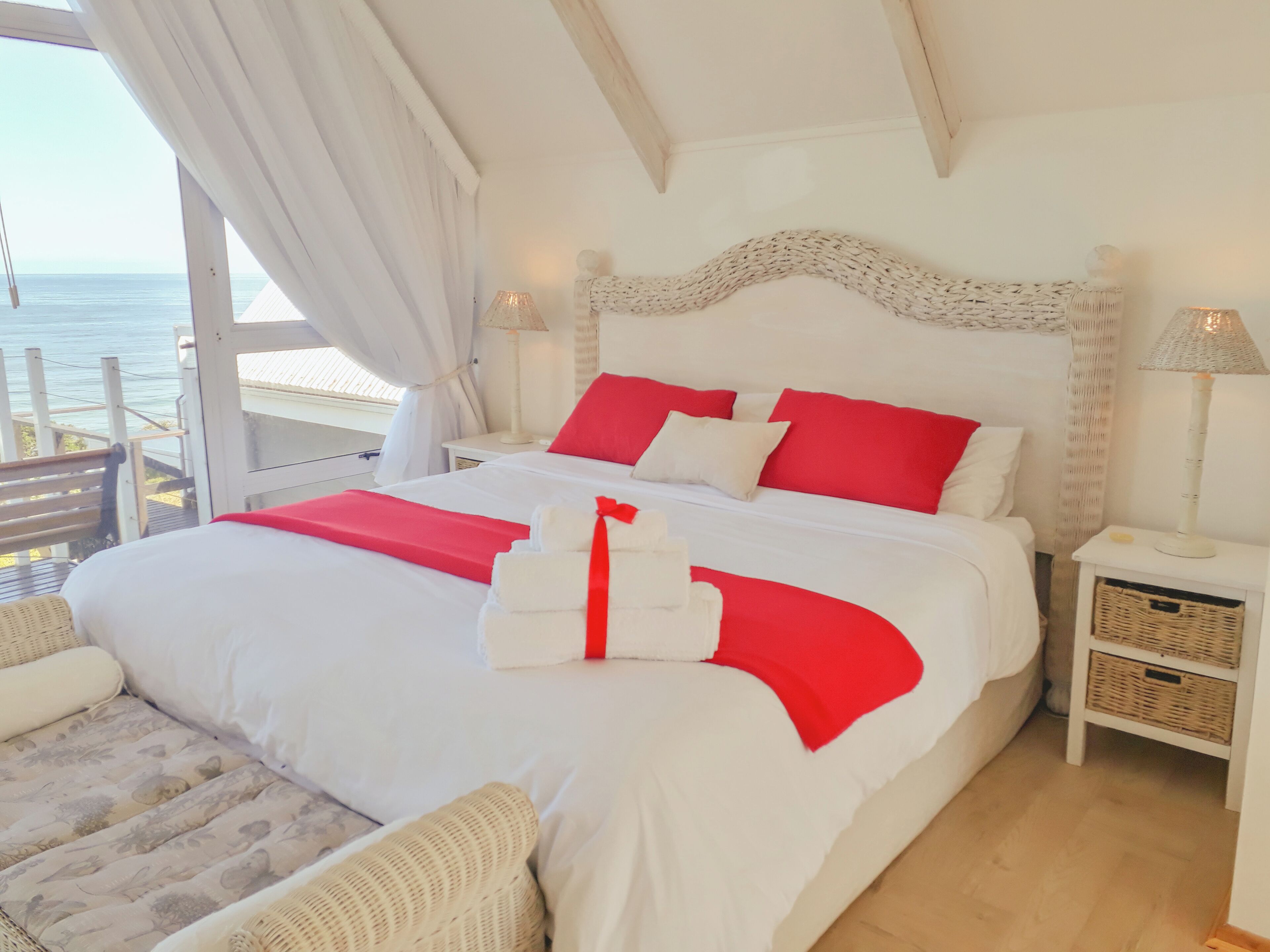 A bedroom with a large bed, red and white bedding, a view of the sea, and a balcony.