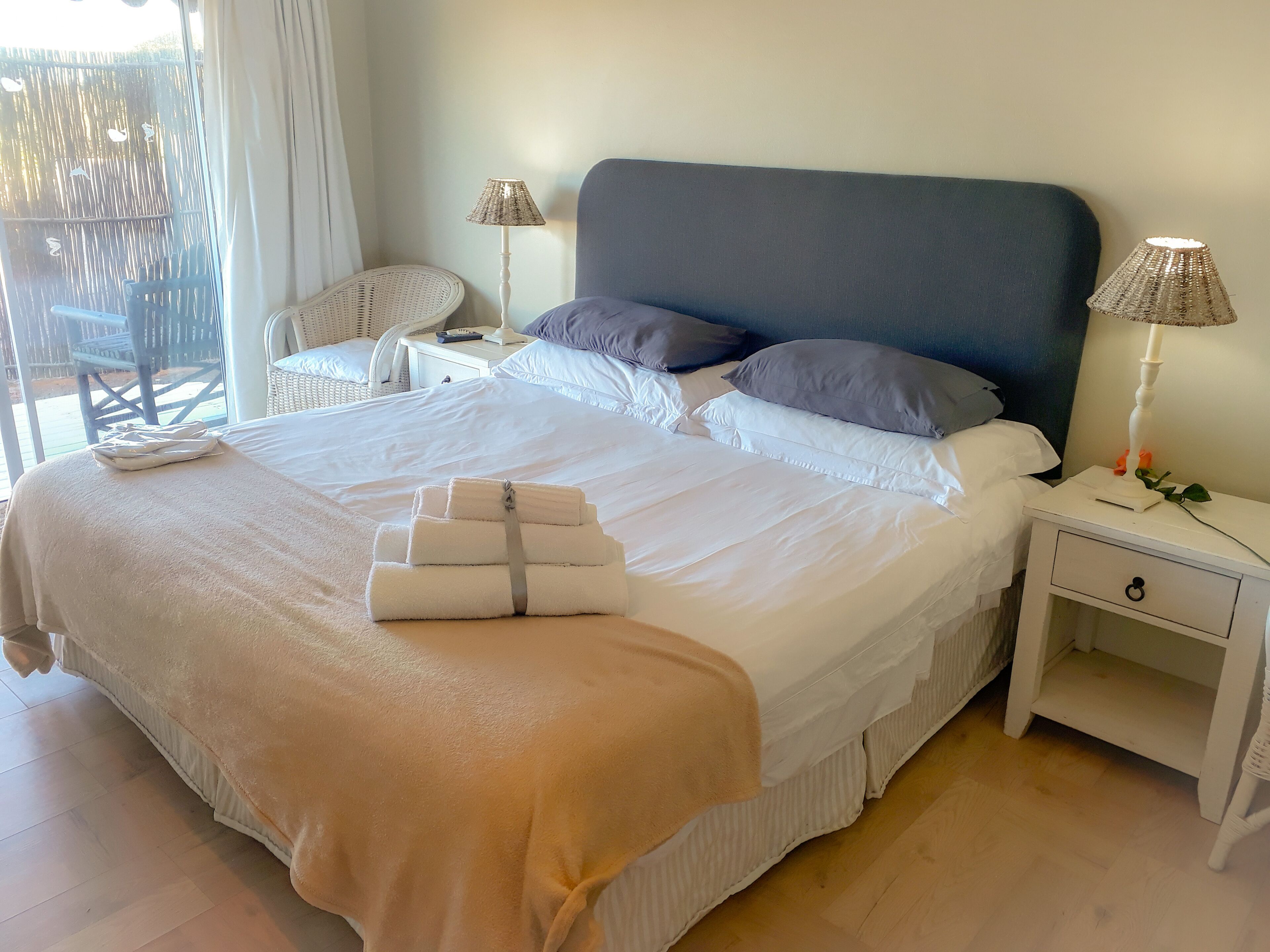A neatly made bed with white linens and a beige throw blanket, two bedside lamps, a white nightstand, and a wicker chair.
