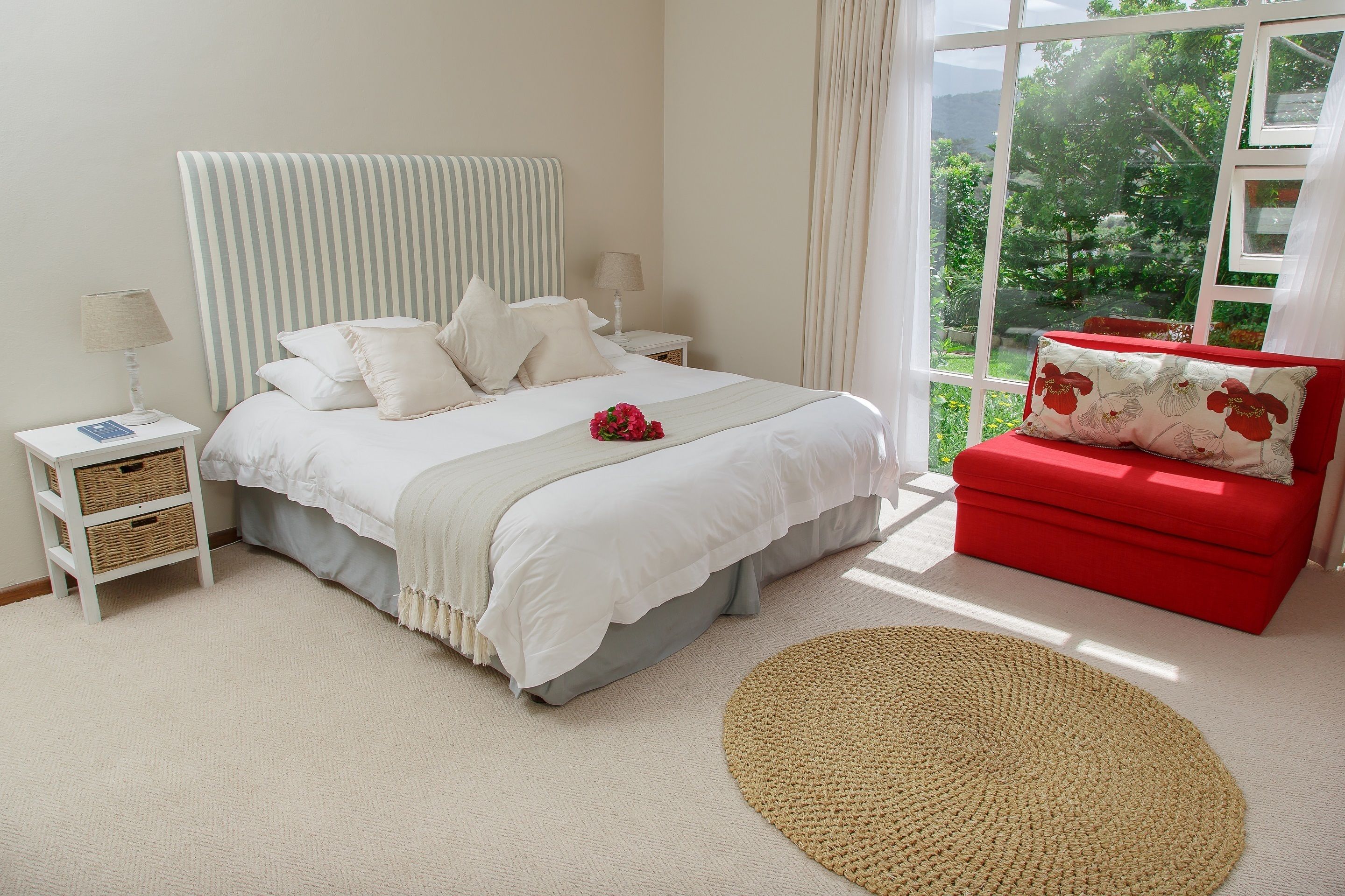 A bedroom with a bed, bedside tables, a red sofa, and a view of greenery through a window.