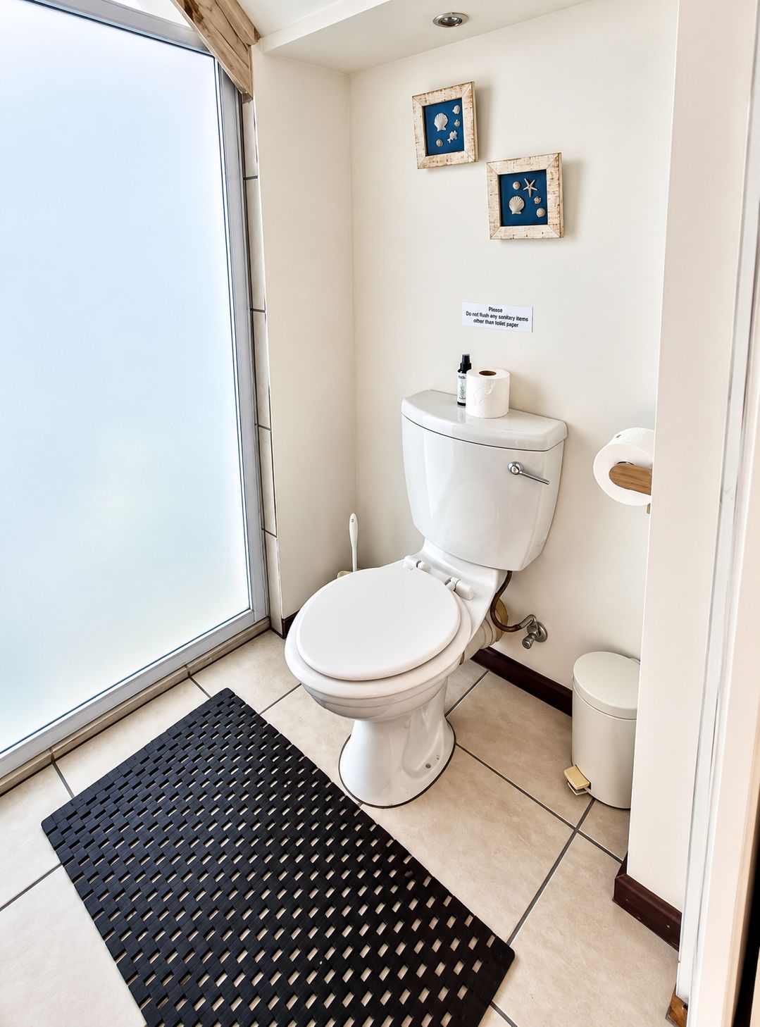 Une petite salle de bain propre, équipée de toilettes, d’un bidet et d’une petite poubelle. Il y a un tapis noir sur le sol et deux tableaux encadrés au mur.