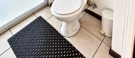 Une petite salle de bain propre, équipée de toilettes, d’un bidet et d’une petite poubelle. Il y a un tapis noir sur le sol et deux tableaux encadrés au mur.