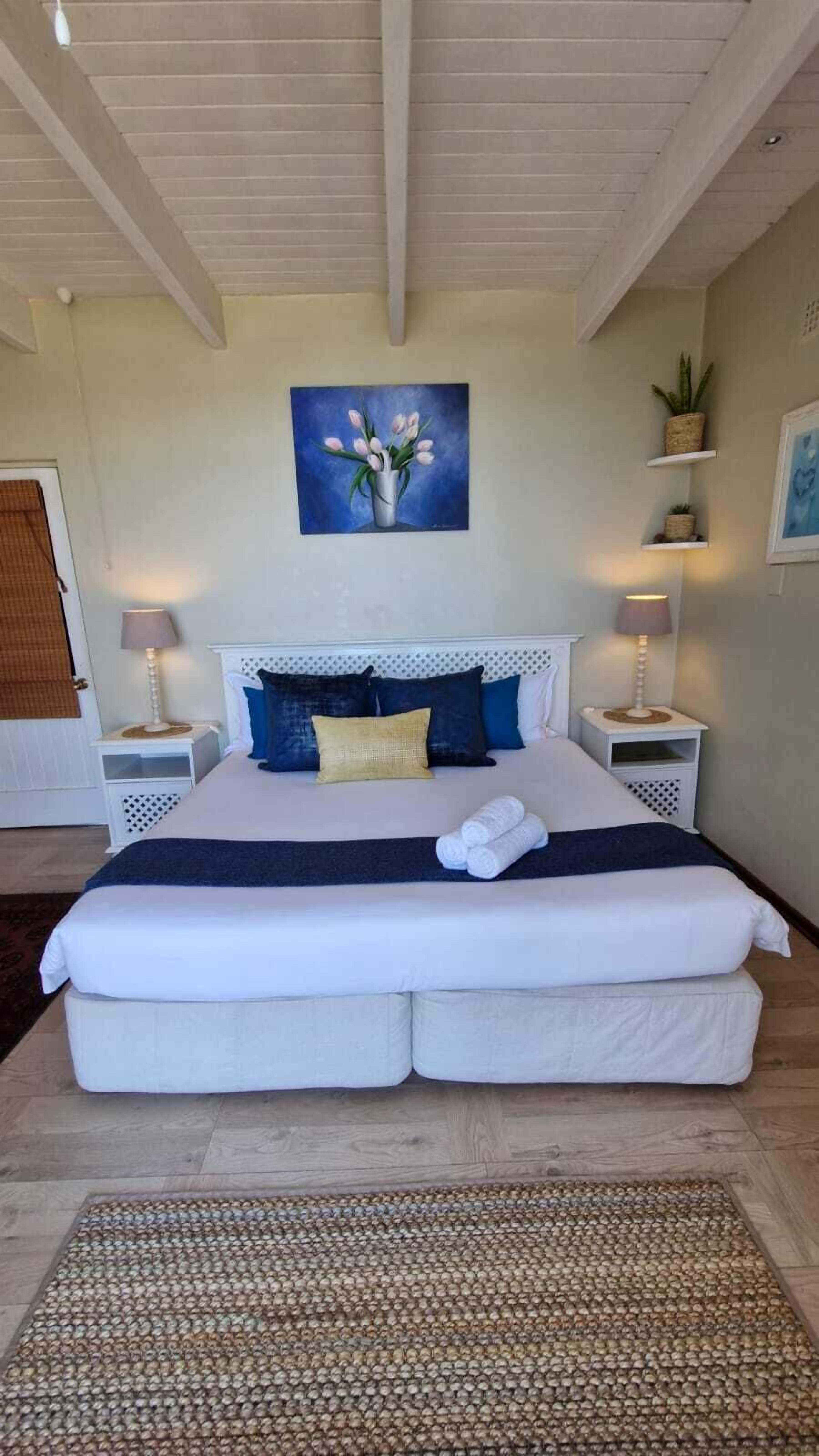 Ein Schlafzimmer mit einem großen Bett, zwei Nachttischen, einem Blumenbild und einer Topfpflanze auf einem Regal.