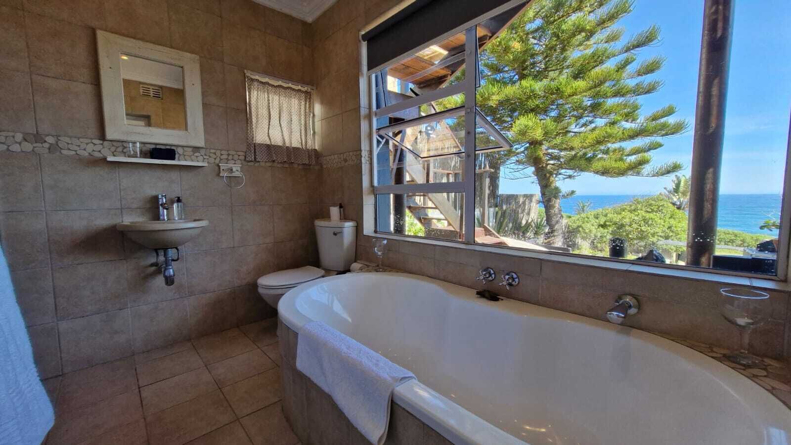 A bathroom with a bathtub, sink, and toilet, with a view of the ocean and trees through a window.