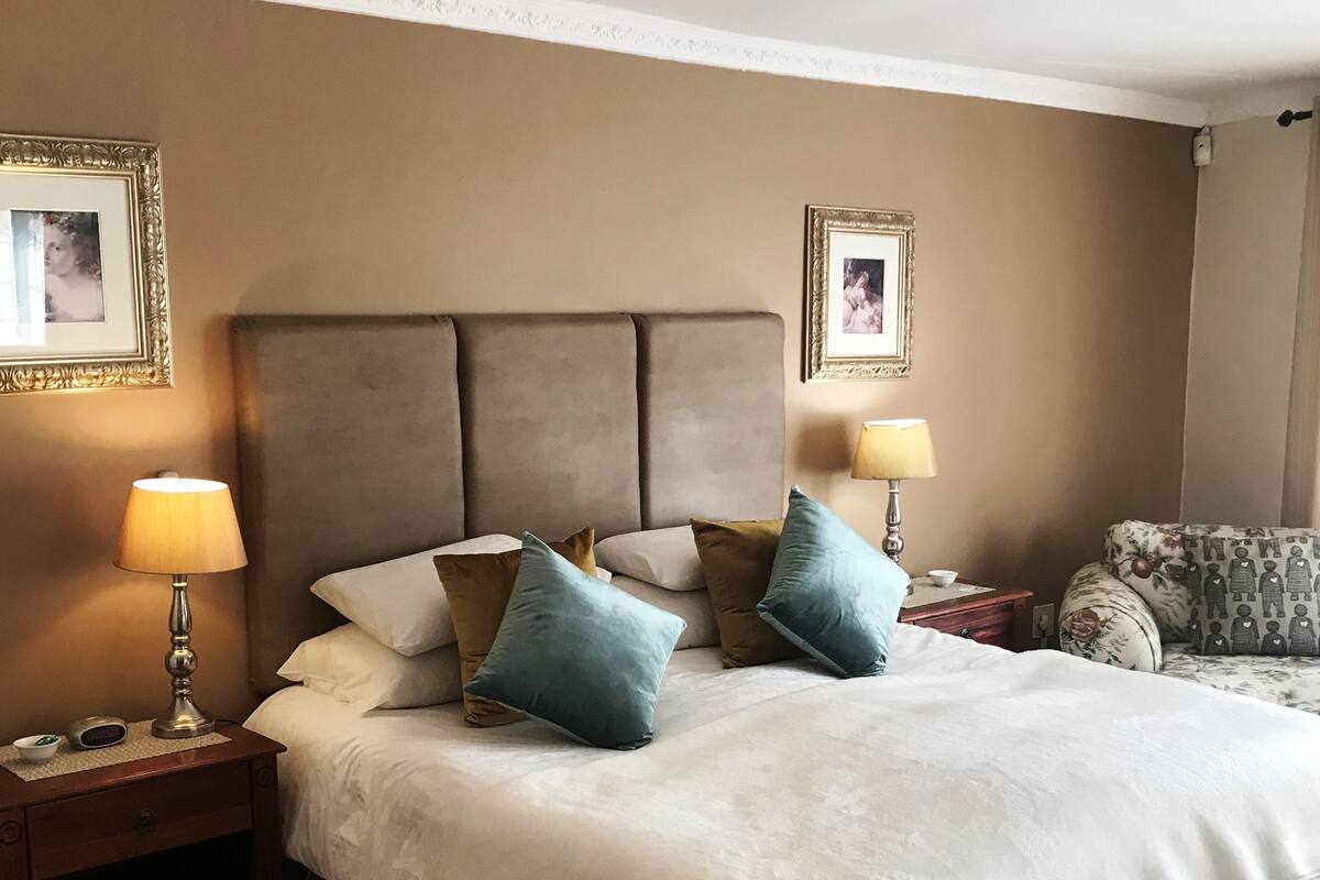 A hotel room with a large bed, two bedside tables with lamps, a floral-patterned armchair, and framed pictures on the walls.