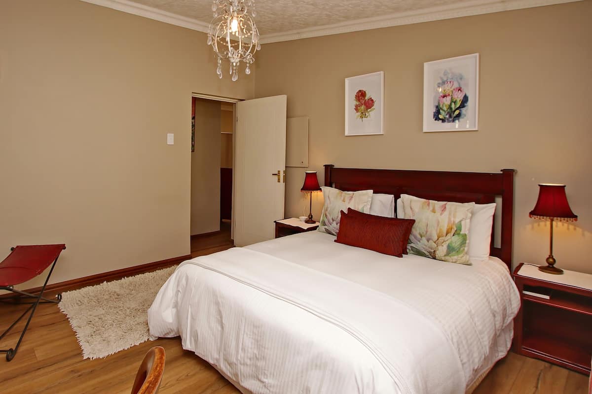 A bedroom with a large bed, two bedside lamps, a chandelier, a red chair, and a door leading to another room.