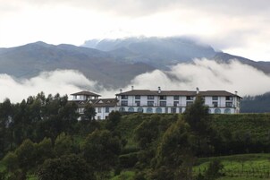 Un edificio grande de varios pisos con fachada simétrica, rodeado de exuberante vegetación y montañas al fondo.