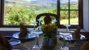 Una mesa de comedor preparada con platos, vasos y un florero, con una vista panorámica de montañas a través de amplias ventanas.