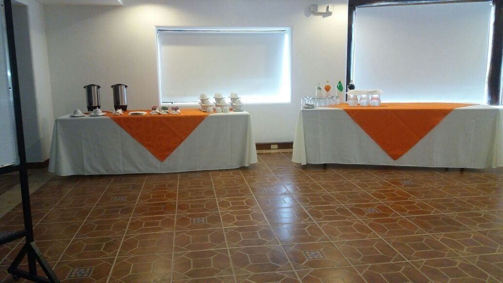 A conference room with two tables, each covered with a white tablecloth and an orange runner. There are two large projection screens on the walls, and the room has a tiled floor.