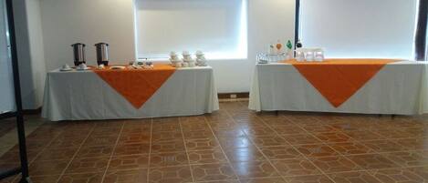 A conference room with two tables, each covered with a white tablecloth and an orange runner. There are two large projection screens on the walls, and the room has a tiled floor.