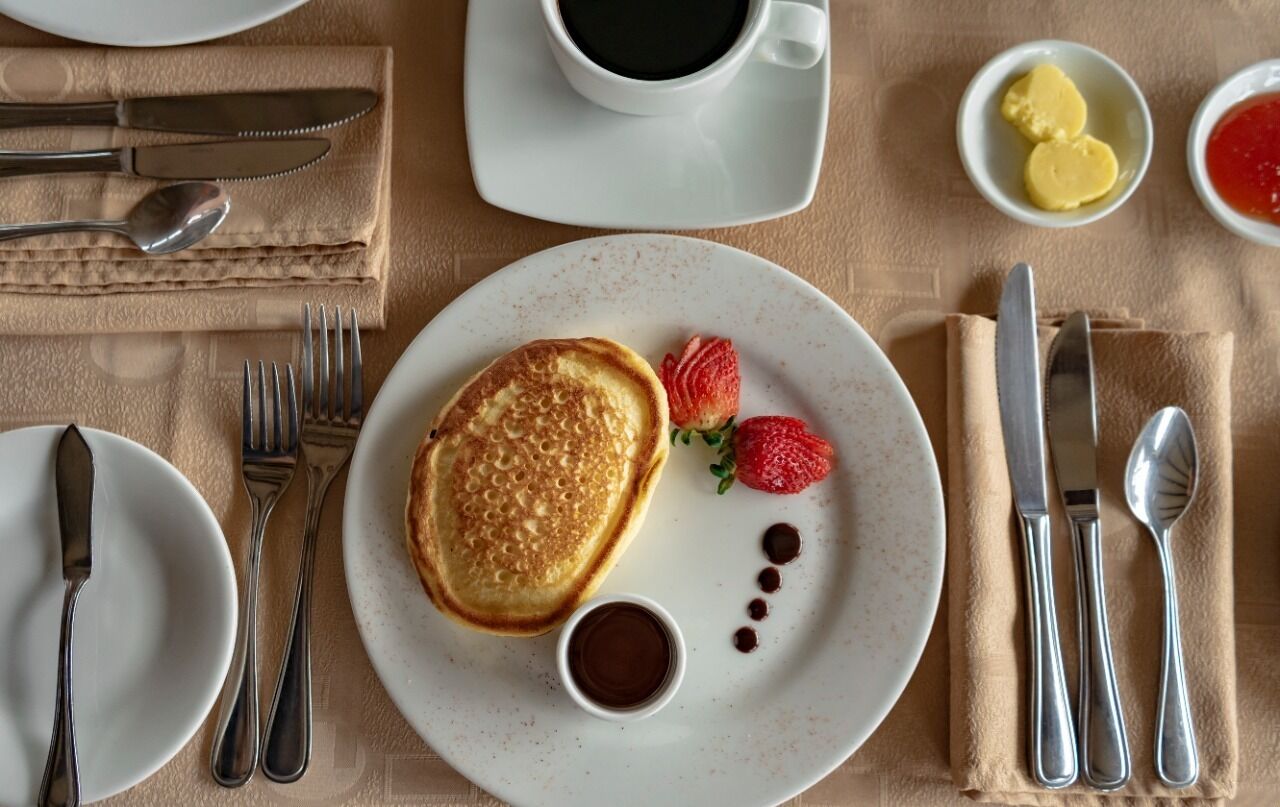 A breakfast setting with pancakes, strawberries, and syrup on a plate, accompanied by cutlery and a cup of coffee.