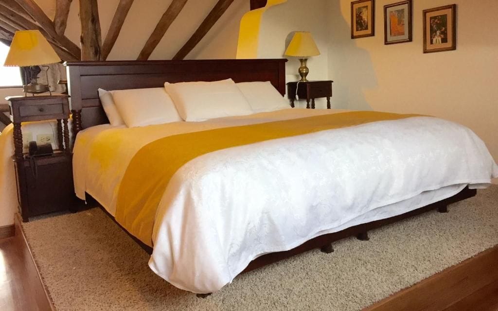 Una cama con colcha blanca y amarilla, dos mesitas de noche con lámparas y un cabecero de madera.